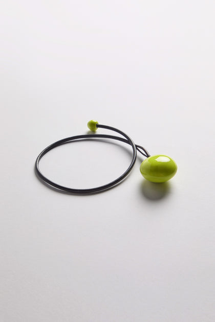 The Handmade Ceramic Bead Necklace features a black cord with a large, glossy, oval green bead with a sculptural twist and a smaller green bead, shown on a white background.