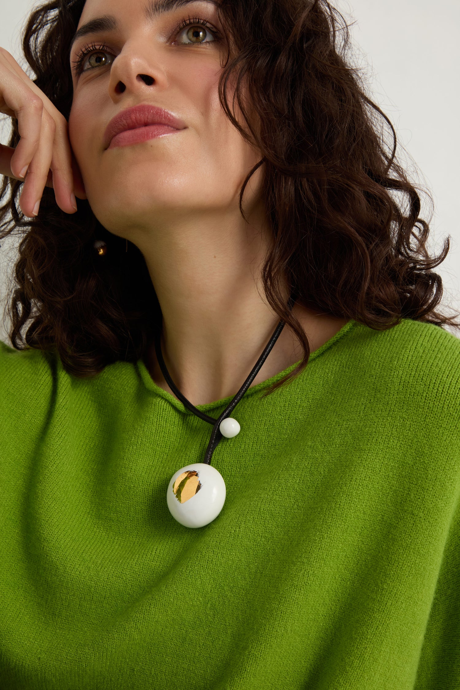 A woman wearing a bright green top and the Handmade Gold Brushstroke Ceramic Bead Necklace, featuring ceramic beads with gold brushstrokes and a round white pendant on a black cord, looks slightly upward.