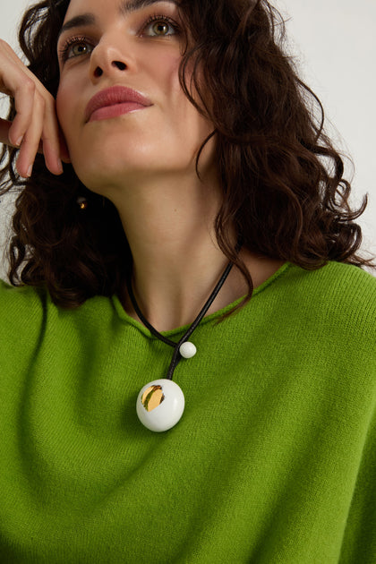 A woman wearing a bright green top and the Handmade Gold Brushstroke Ceramic Bead Necklace, featuring ceramic beads with gold brushstrokes and a round white pendant on a black cord, looks slightly upward.