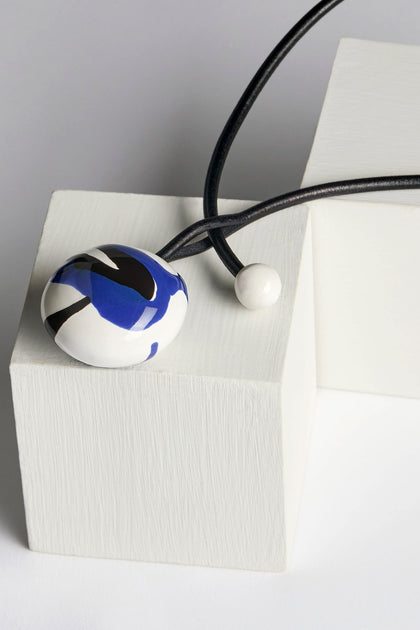 The Handmade Ceramic Painted Bead Necklace, featuring a spherical pendant adorned with a blue, black, and white pattern on a black cord, is elegantly displayed on a textured white cube, highlighting the traditional craftsmanship of its design.