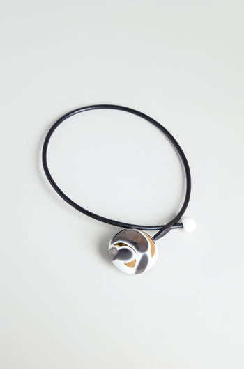 The Slate Handmade Ceramic Painted Bead Necklace features a black cord, metallic abstract pendant, and white ceramic bead—blending French-inspired elegance with artisan craftsmanship. Displayed on a light surface for a modern look.