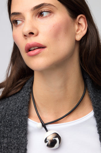 A woman with long dark hair wears a white shirt, a dark textured cardigan, and the Slate Handmade Ceramic Painted Bead Necklace—a striking artisan piece featuring a large metallic pendant.