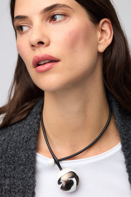 A woman with long dark hair wears a white shirt, a dark textured cardigan, and the Slate Handmade Ceramic Painted Bead Necklace—a striking artisan piece featuring a large metallic pendant.