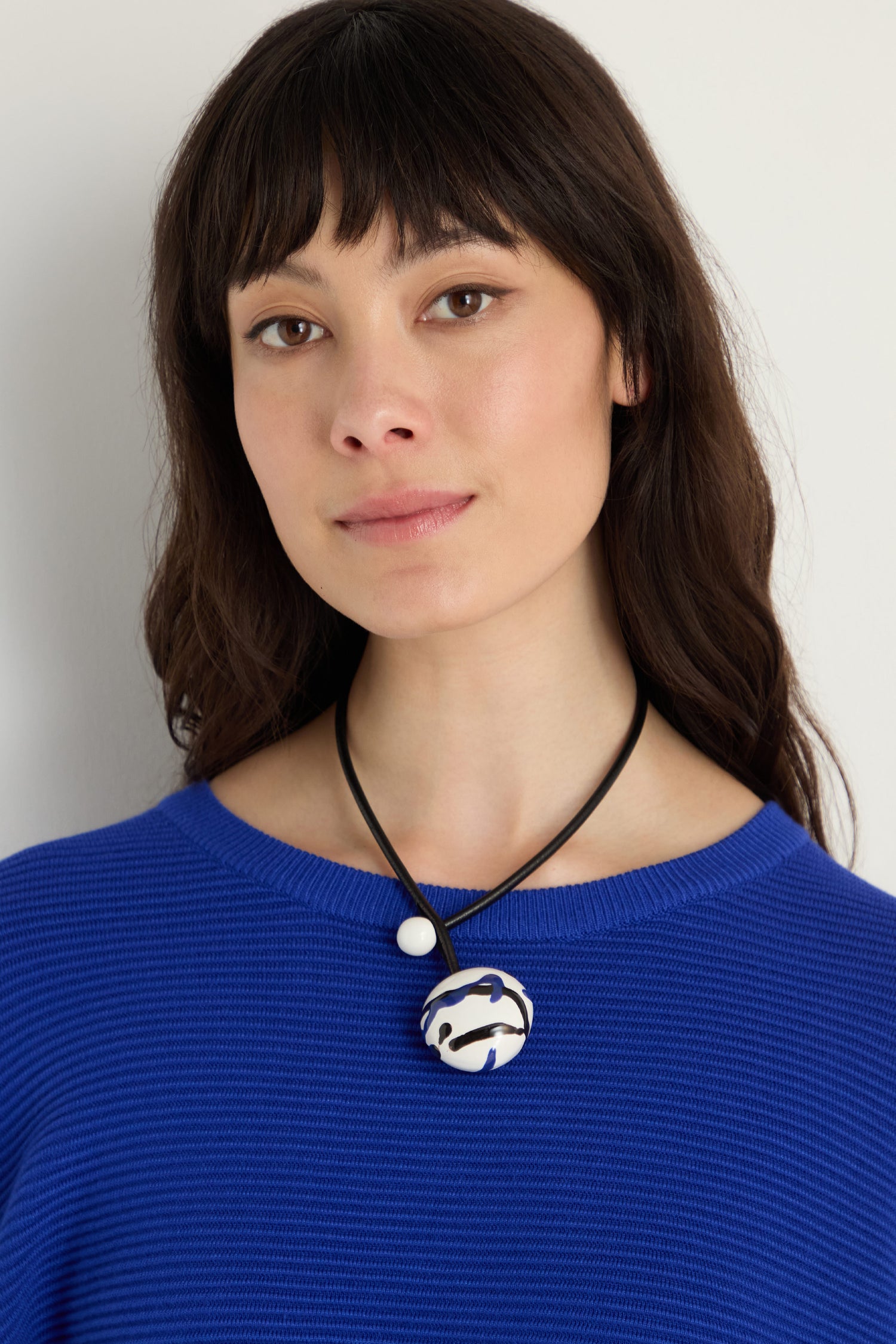 A woman with long dark hair wears a blue textured sweater and the Handmade Painted Ceramic Bead Necklace, featuring a round, abstract painted bead on a black leather cord, standing against a plain light background.