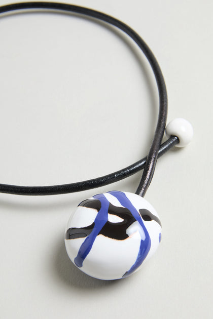 The Handmade Painted Ceramic Bead Necklace features a black leather cord, a round white pendant with blue and black abstract lines, and is accented by a small painted ceramic bead.