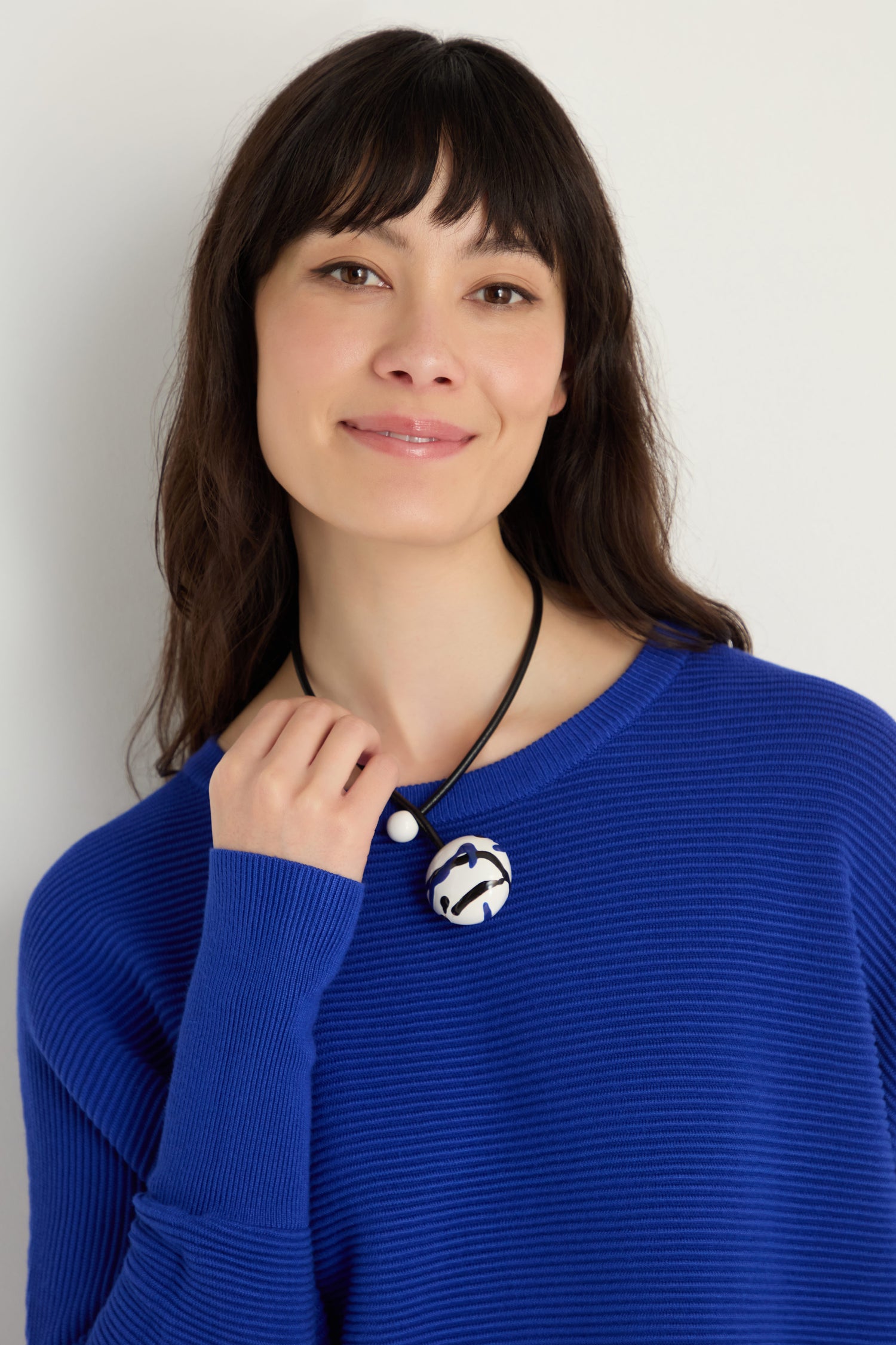Woman with straight dark hair wears a bright blue ribbed sweater and the Handmade Painted Ceramic Bead Necklace, which features a round painted ceramic bead on a black leather cord, standing against a light background.