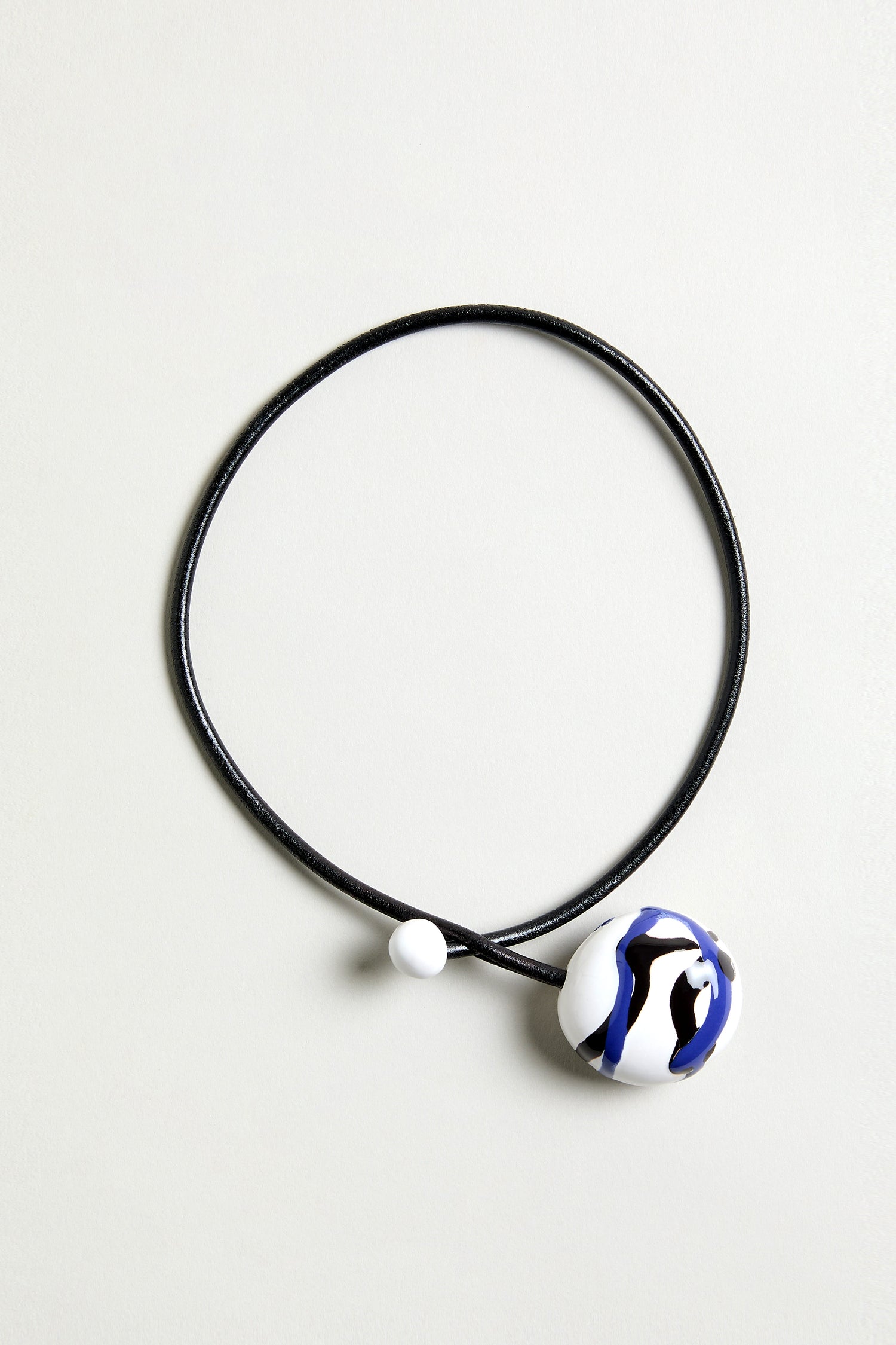 The Handmade Painted Ceramic Bead Necklace features a black leather cord, a round white pendant with blue and black abstract patterns, and a small hand-painted ceramic bead.