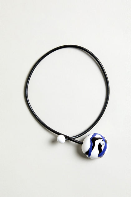 The Handmade Painted Ceramic Bead Necklace features a black leather cord, a round white pendant with blue and black abstract patterns, and a small hand-painted ceramic bead.