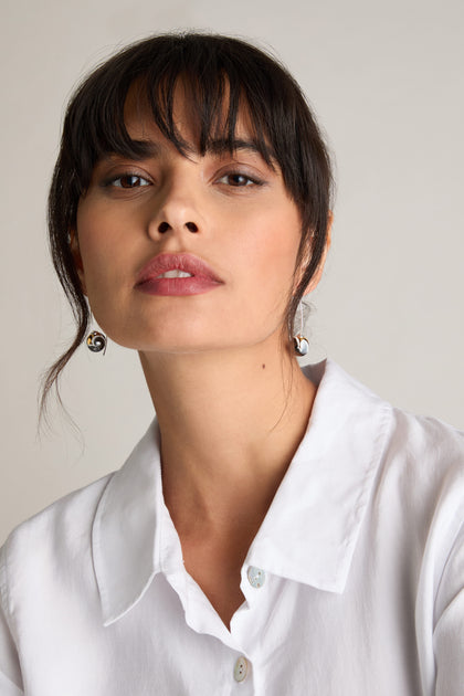 A person with dark hair and bangs is wearing a white collared shirt and Handmade Ceramic Graphic Earrings, which feature geometric shapes. They have a neutral expression against a light background, showcasing a modern minimalist style.