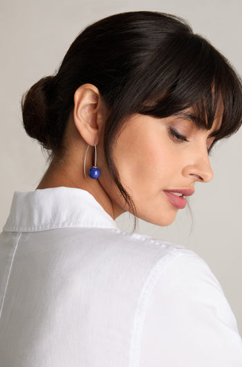 A woman with dark hair in a low bun is wearing Handmade Ceramic Bead Earrings crafted from 925 sterling silver. She is dressed in a white shirt and looking down.