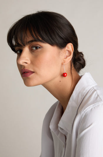 A woman with straight dark hair tied back, wearing a white shirt and Handmade Ceramic Bead Earrings in red paired with 925 sterling silver, looks to the side with a neutral expression.