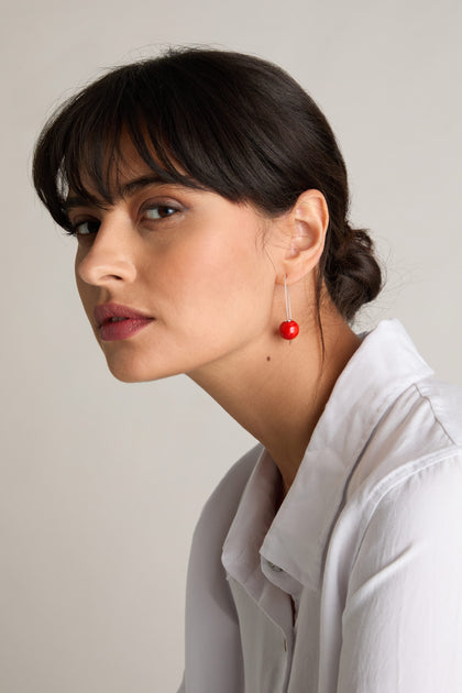 A woman with straight dark hair tied back, wearing a white shirt and Handmade Ceramic Bead Earrings in red paired with 925 sterling silver, looks to the side with a neutral expression.