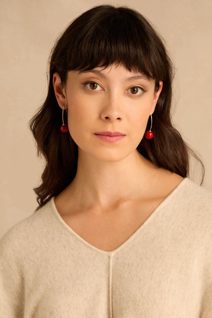 A person wearing Handmade Deep Red Ceramic Bead Earrings, featuring long silver wires with a deep red ceramic bead at the end of each.