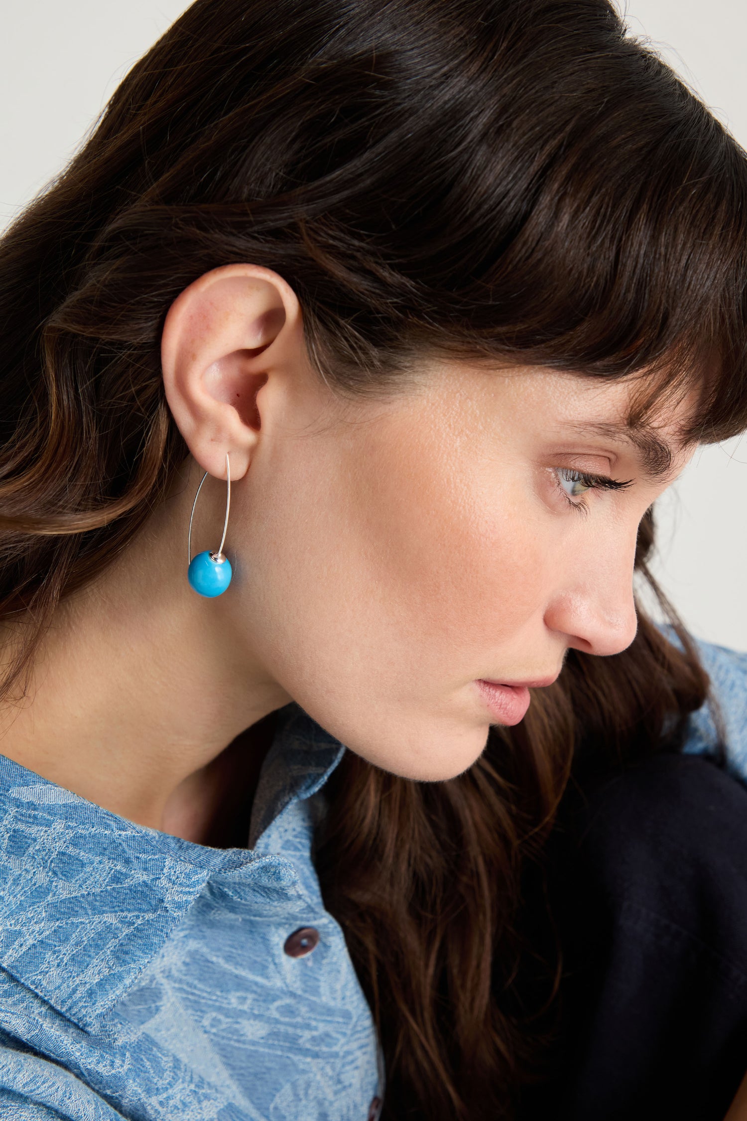 A woman with long brown hair is shown in side profile wearing Handmade Ceramic Bead Earrings with bright turquoise beads on silver hooks and a blue patterned shirt.
