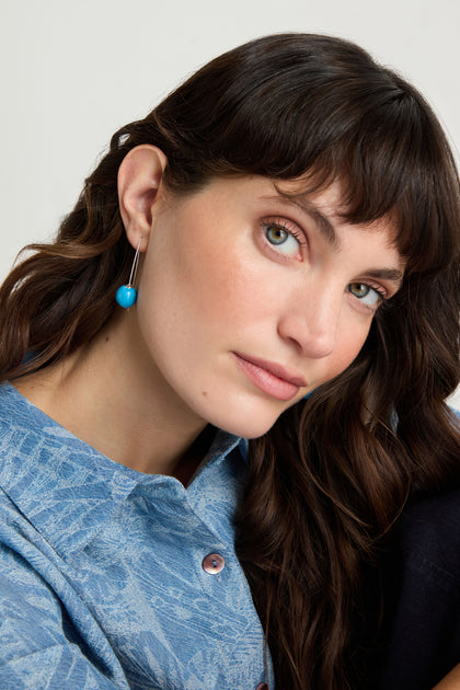 A woman with long brown hair, wearing a blue patterned shirt and the Handmade Ceramic Bead Earrings with silver hooks, looks at the camera against a plain background.