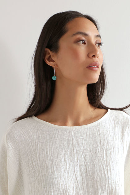 A woman with long dark hair wears a textured white top and Handmade Ceramic Bead Earrings with an organic finish, looking slightly right against a plain light background.
