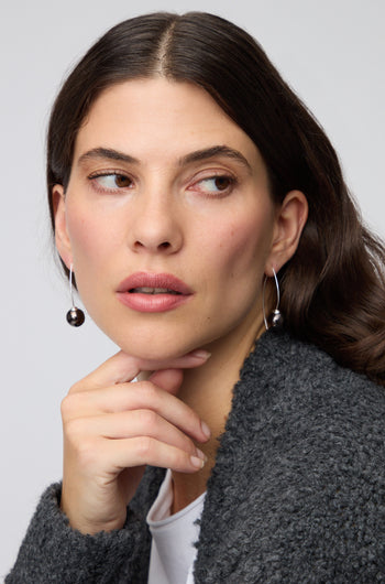 A woman with long brown hair wears Handmade Ceramic Graphic Metal Earrings and a textured dark gray jacket, resting her chin on her hand and gazing thoughtfully to the side.