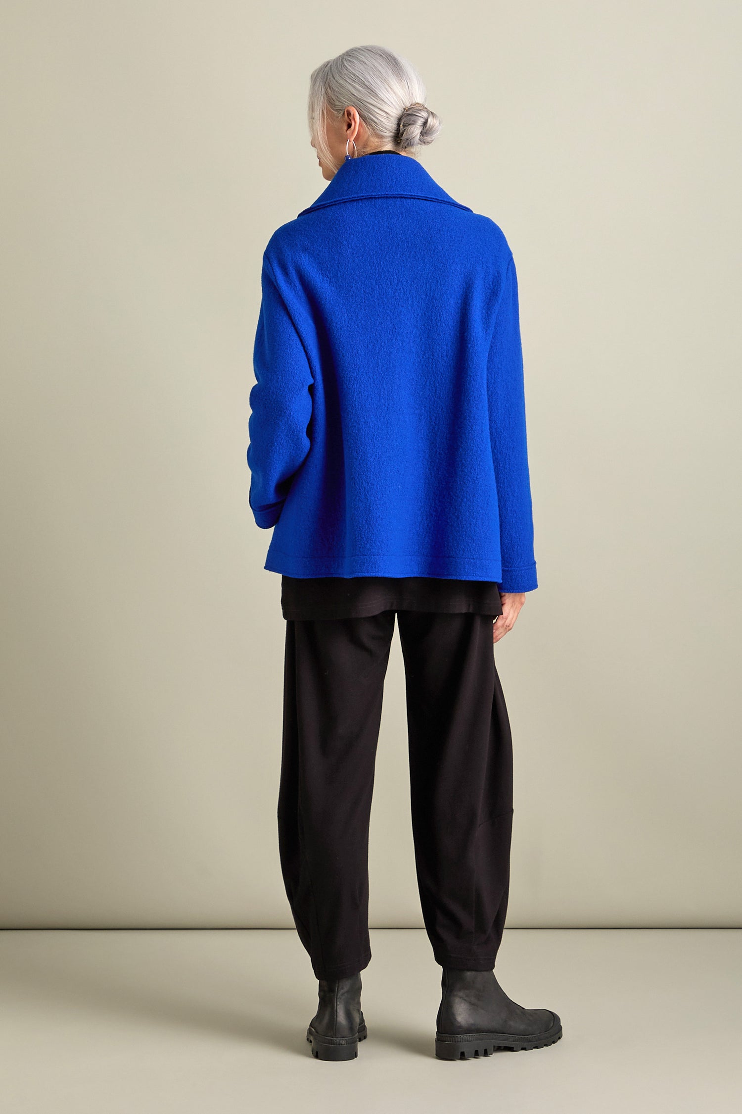 A person with gray hair, styled in a bun, is wearing a royal blue Boiled Wool Zip Boxy Jacket along with black pants and black boots. They are standing with their back to the camera against a plain background.