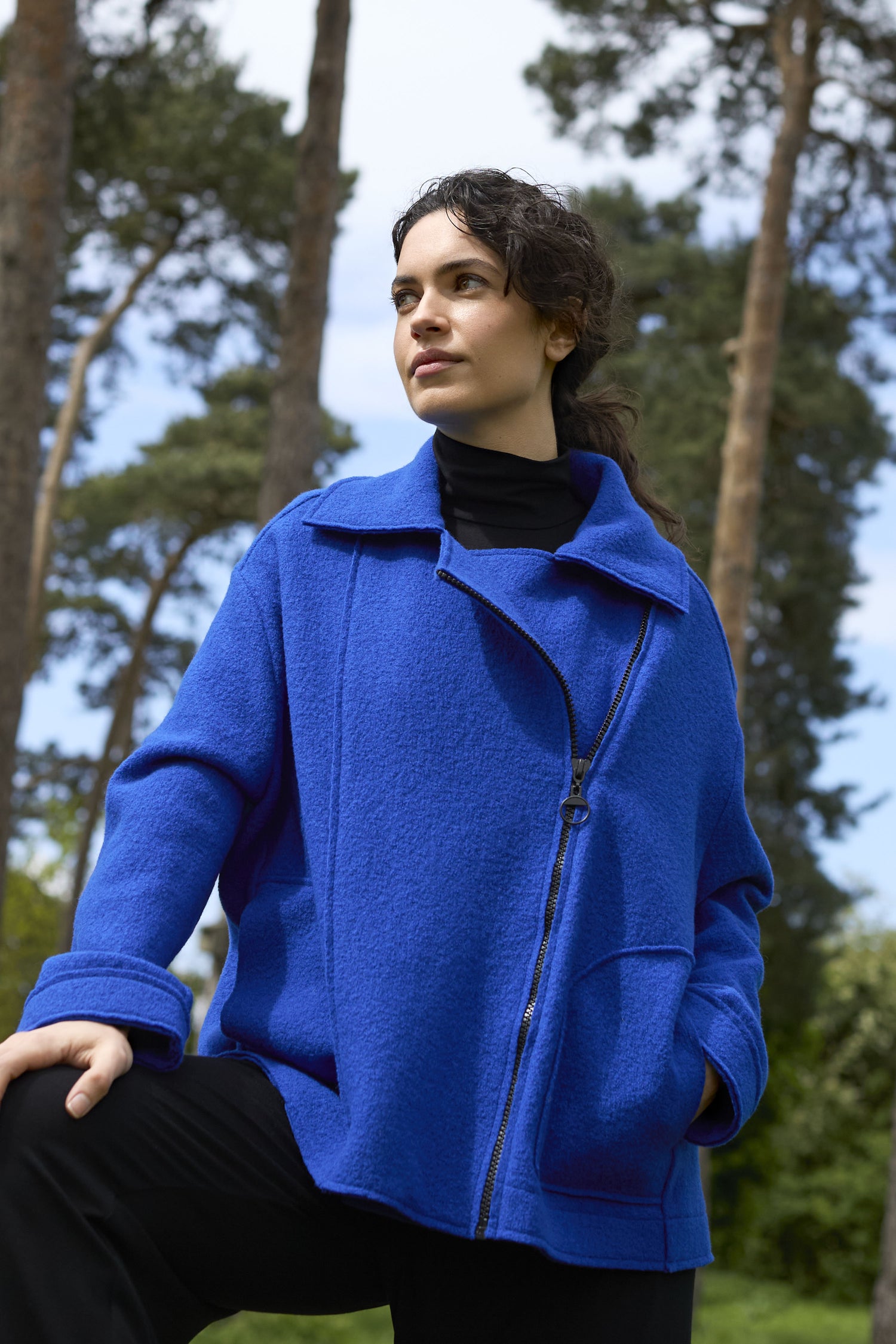 A person wearing a Boiled Wool Zip Boxy Jacket in royal blue stands outdoors among trees, looking off into the distance.