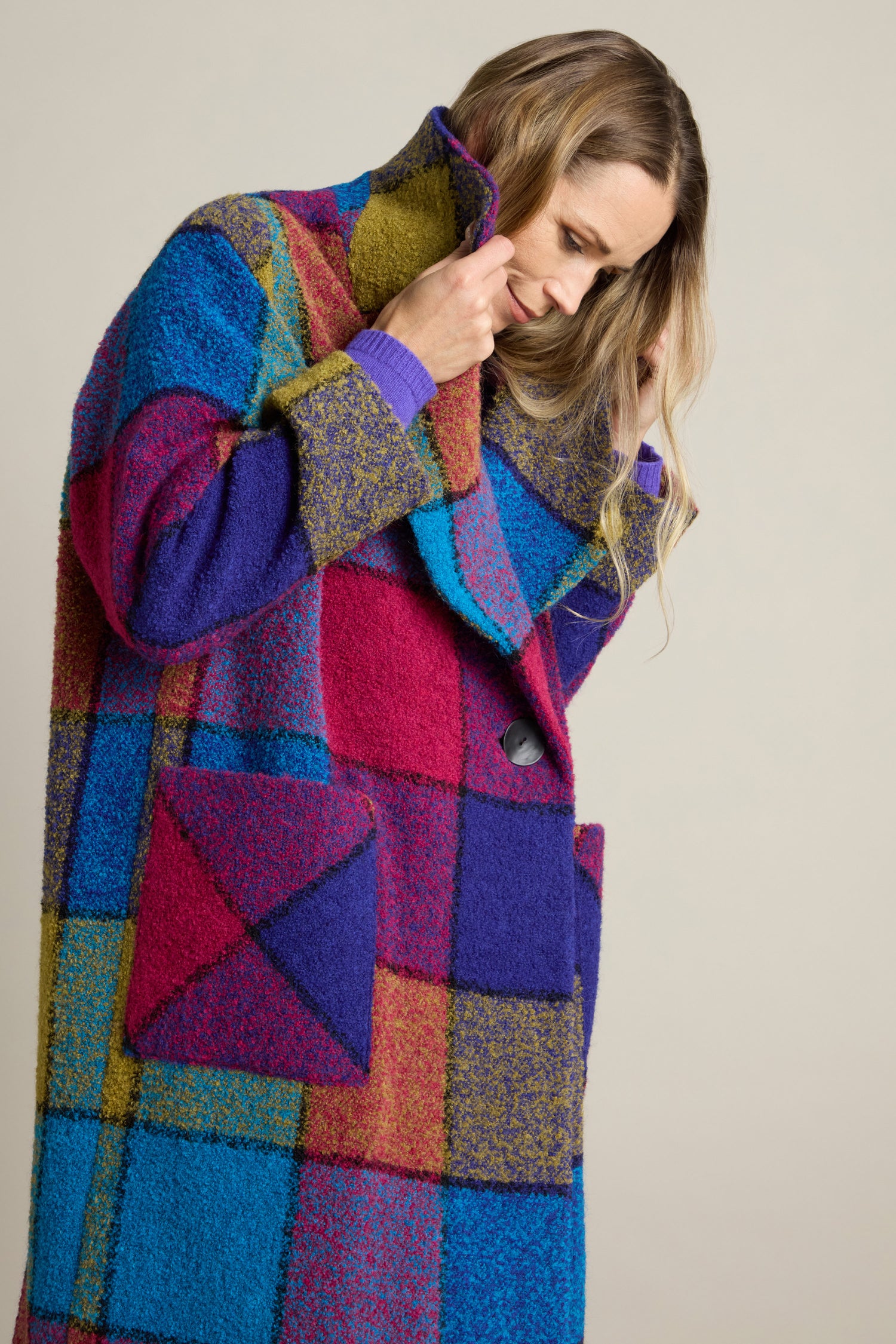 An individual is showcasing a vibrant statement piece by donning the Check Wool Coat, adorned with multicolored patchwork squares in shades of blue, red, purple, and yellow. The coat features large pockets and they are seen adjusting the collar against a plain neutral background.