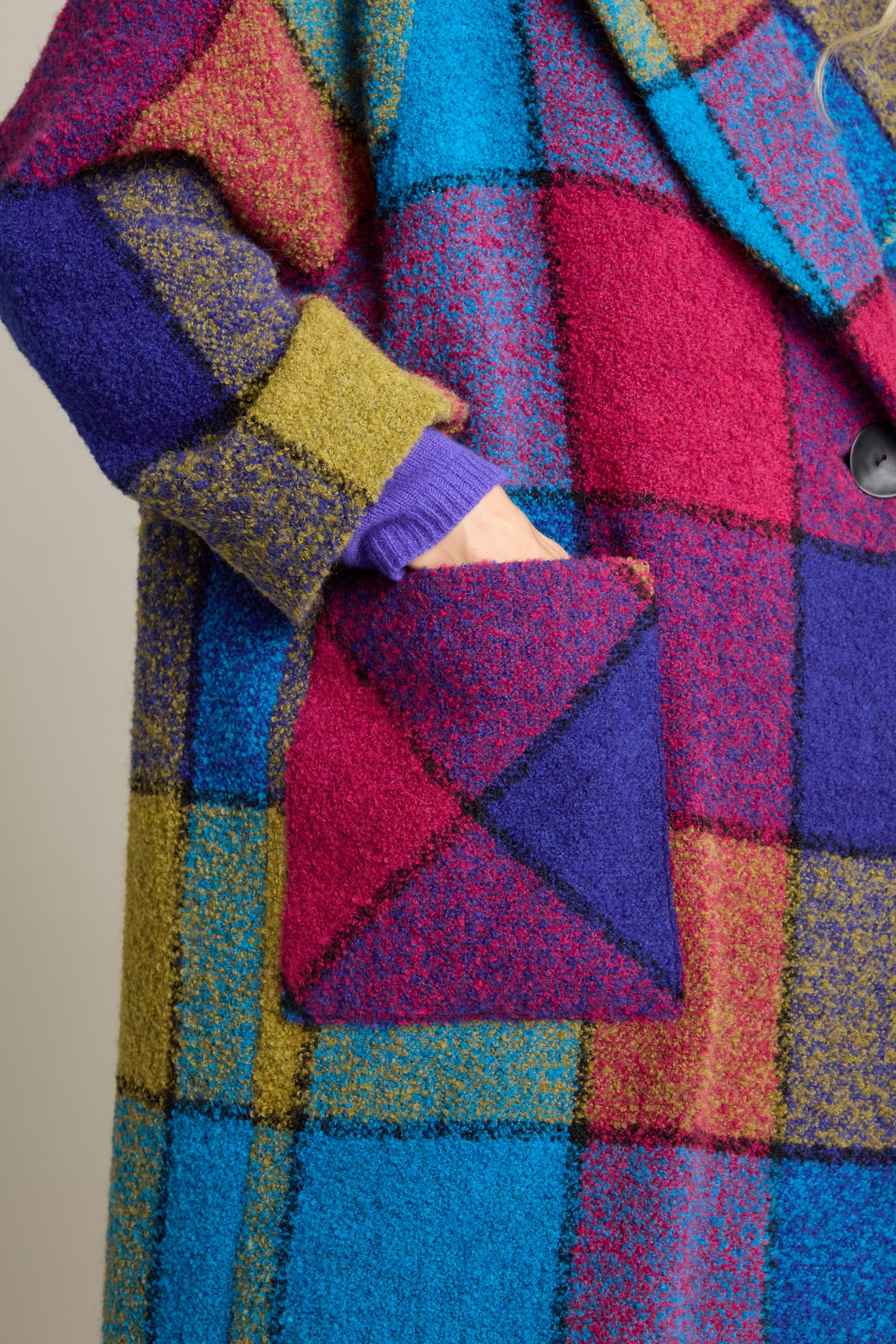 Close-up of a person’s hand in the pocket of the Check Wool Coat made from a boucle wool blend featuring a colorful, patchwork pattern with shades of blue, purple, red, and green—truly a vibrant statement piece.