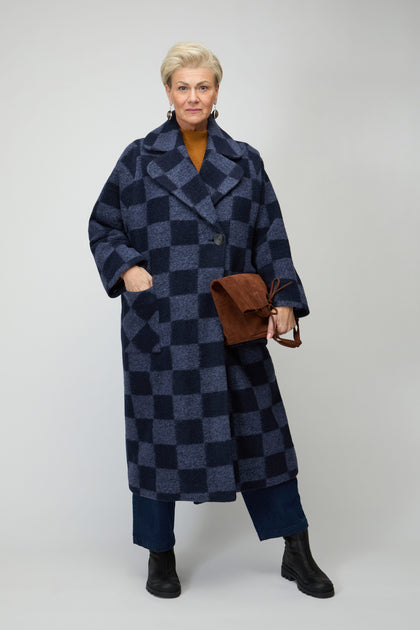 An older woman wears the Checkerboard Wool Coat in blue, paired with blue pants and black boots, holding a brown bag against a plain light background.