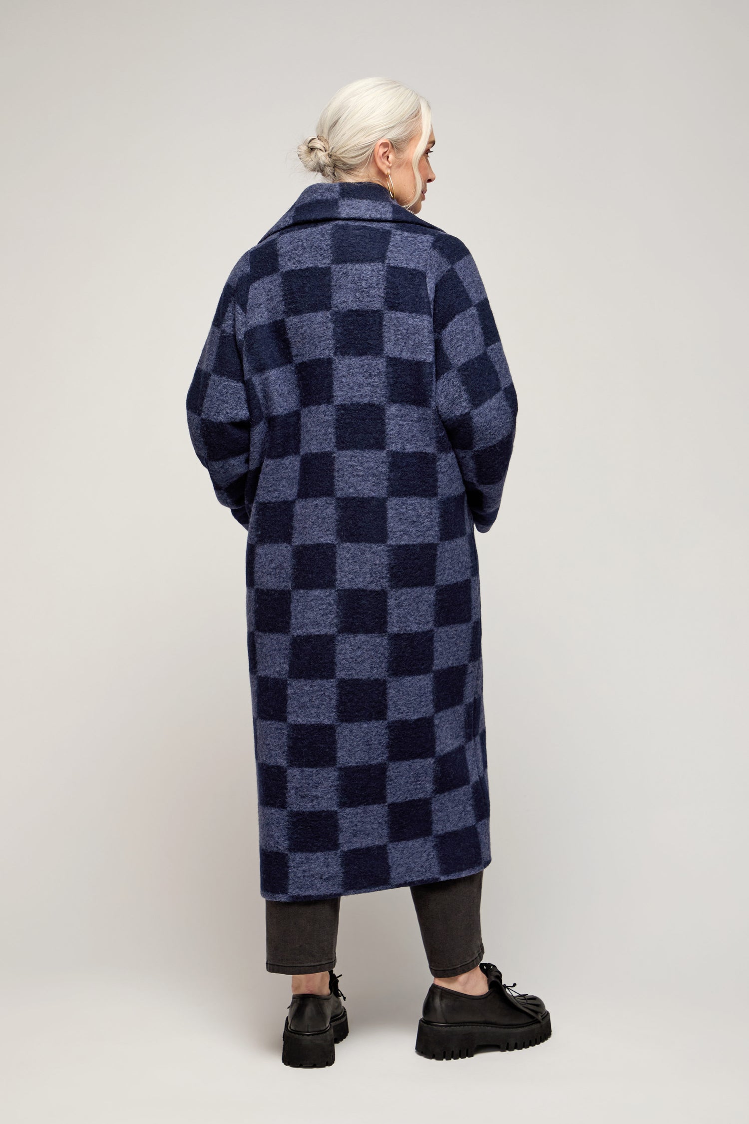 A person with grey hair in a low bun wears the Checkerboard Wool Coat, dark pants, and black shoes, standing with their back to the camera against a plain background.