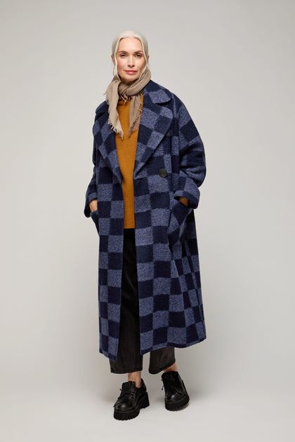 A woman with gray hair in a tan headscarf, brown sweater, black pants, and black shoes stands facing forward against a plain background, wearing the Checkerboard Wool Coat.