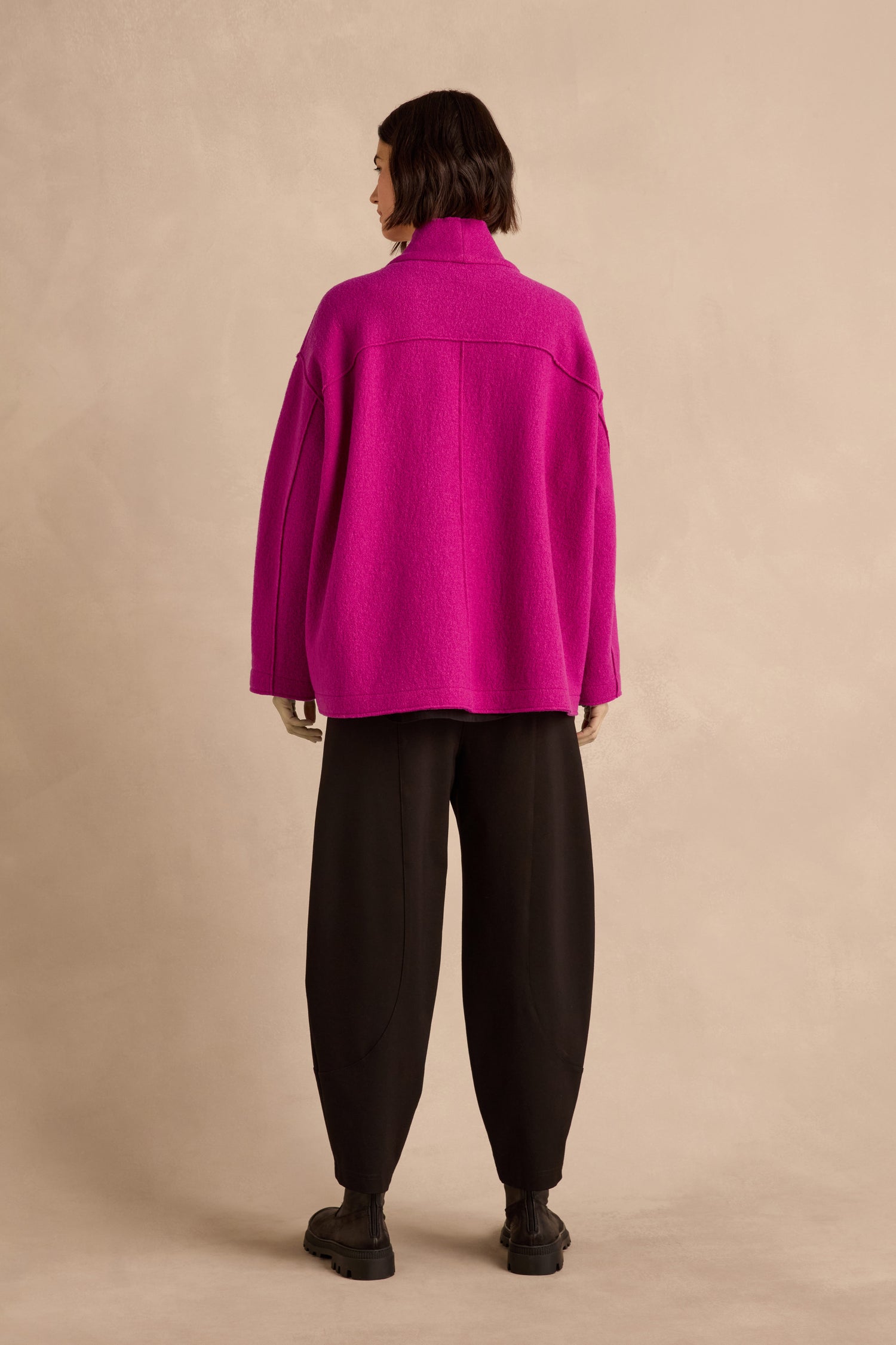 A person stands with their back to the camera, wearing the Magenta Boiled Wool Cocoon Jacket—an oversized piece with a modern silhouette—paired with black wide-leg pants and black shoes against a beige background.