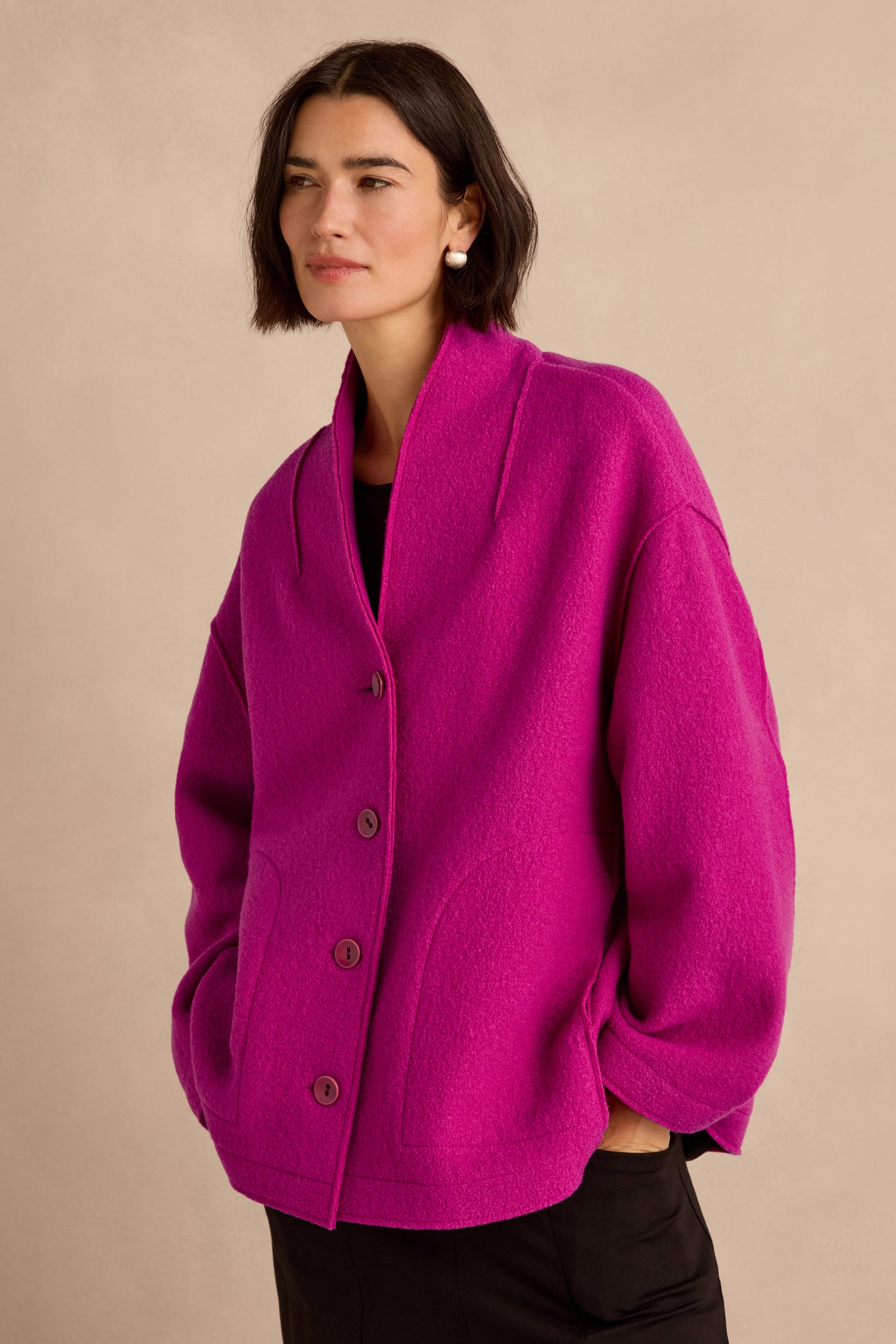 A woman with short dark hair stands against a neutral background, hands in pockets, modeling the Magenta Boiled Wool Cocoon Jacket—a bright jacket made in England and featuring a relaxed cocoon silhouette.