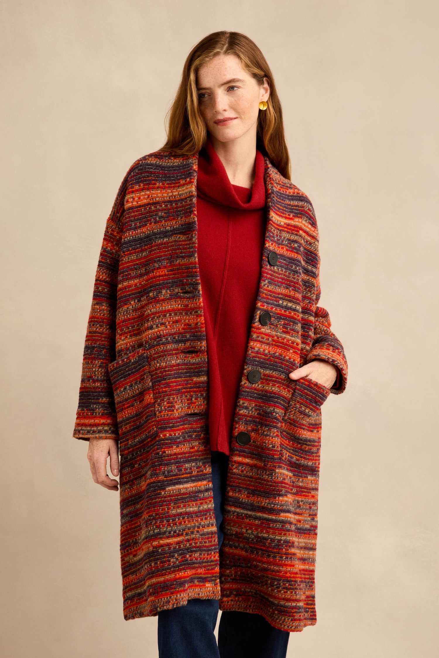A woman stands against a plain beige background, wearing the Artisan Wool Blend Stripe Coat—an oversized coat with long multicolored stripes—layered over a red top, with one hand in her pocket.