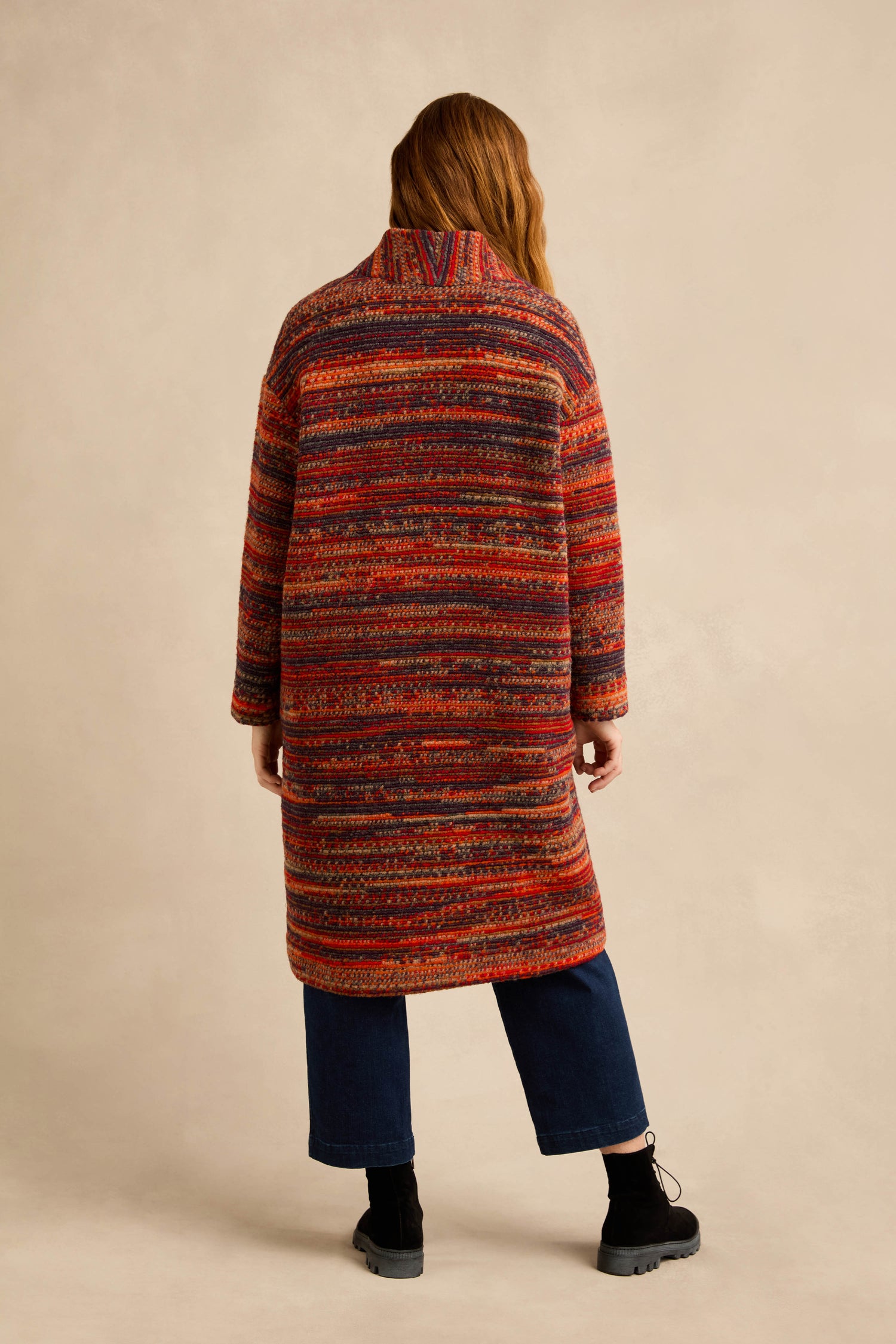 A person with long hair stands with their back to the camera against a beige background, wearing the Artisan Wool Blend Stripe Coat in red, orange, and purple hues. The look is paired with dark cropped jeans and black boots.