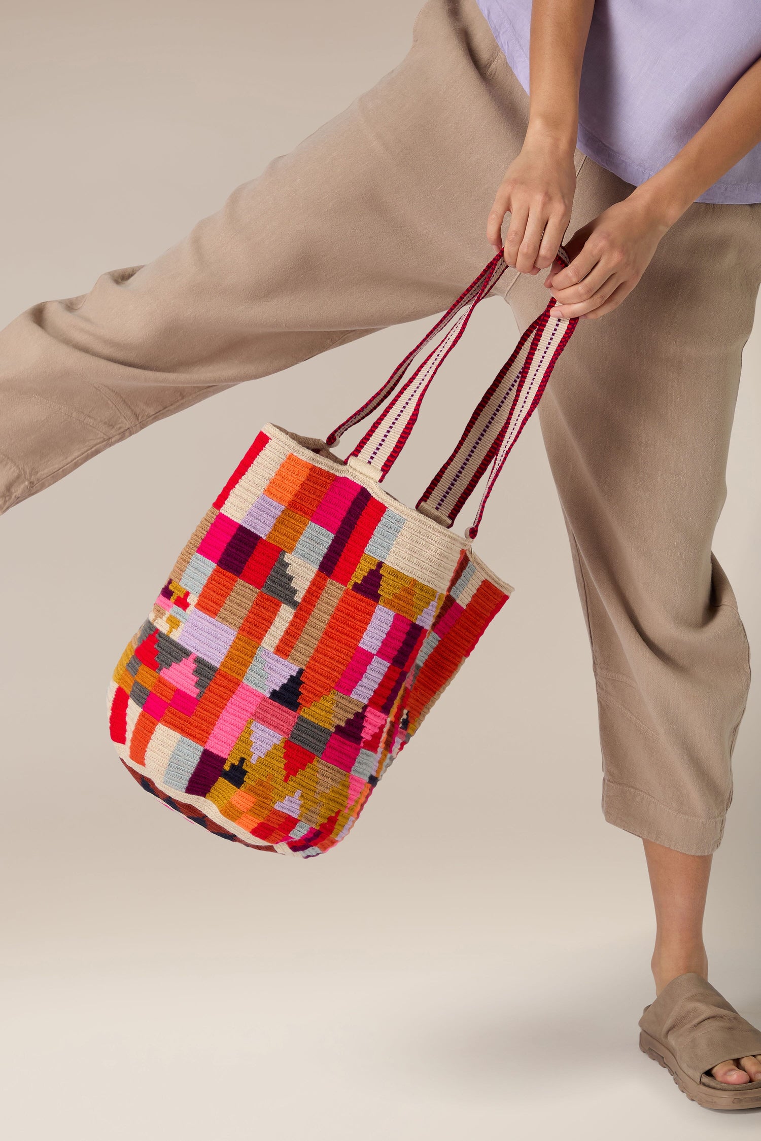 Person holding a colorful, rectangular-patterned woven tote bag with striped handles. This Handcrafted Cala Tote Bag showcases vibrant geometric patterns created by skilled artisans. They are dressed in beige pants, a lavender shirt, and brown shoes. One leg is raised in a playful manner.