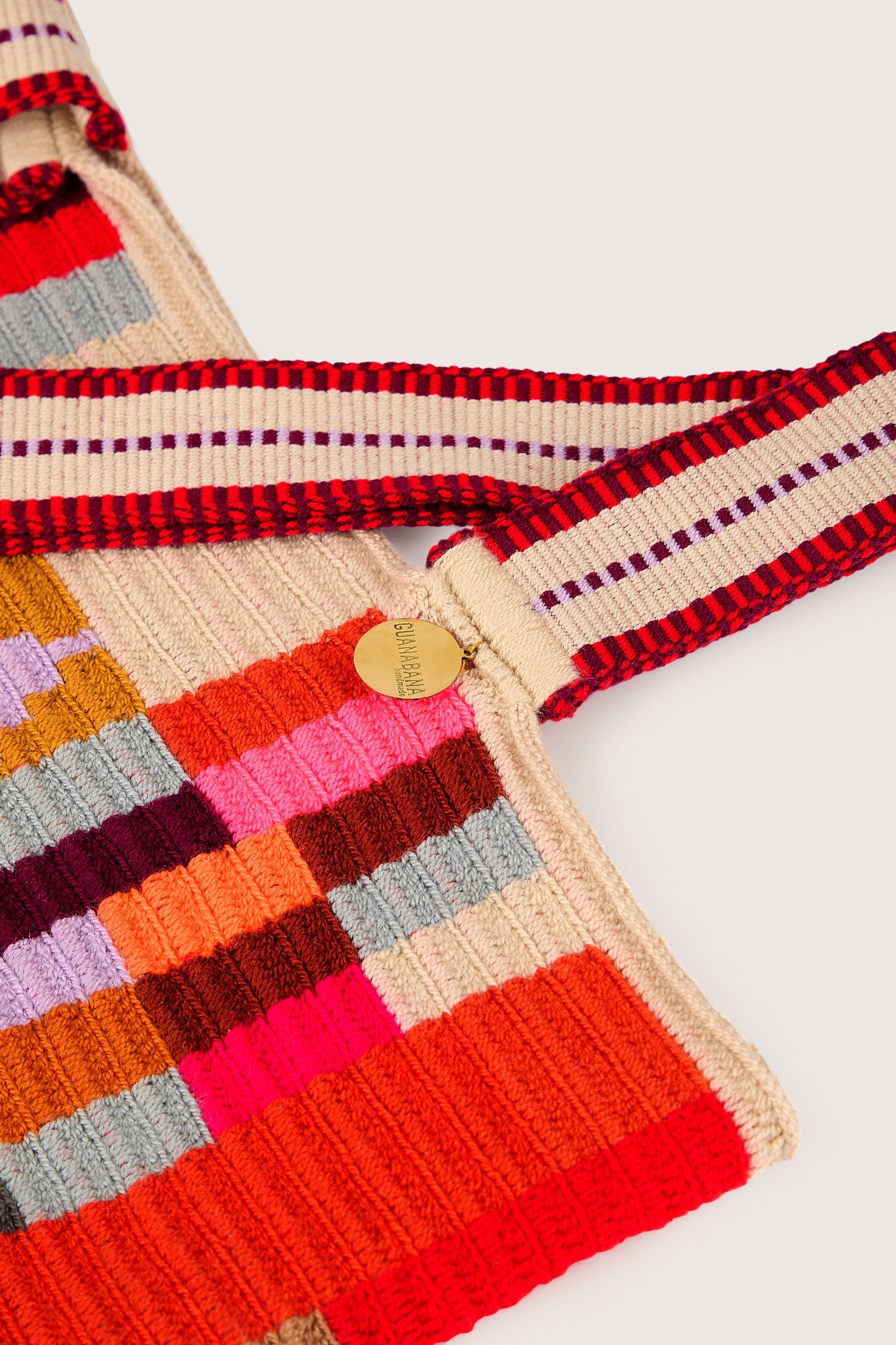 Close-up image of the **Handcrafted Cala Tote Bag**, showcasing geometric patterns in its multicolored knit and complemented by striped red, beige, and purple shoulder straps. A small, round gold-colored tag adds a touch of elegance to this creation by skilled artisans.