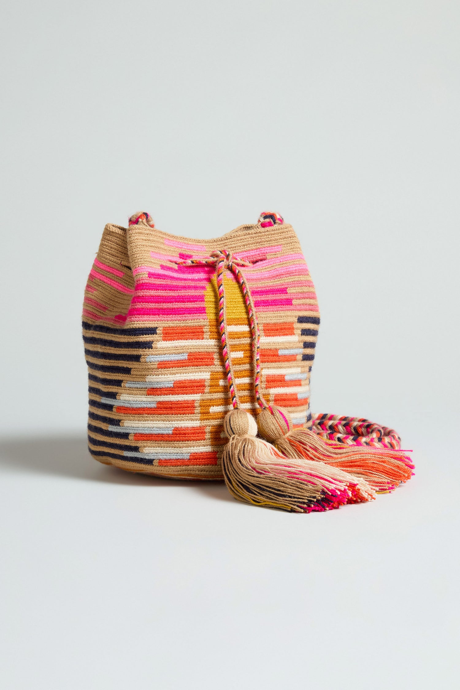 The Handcrafted Crossbody Tassel Bag is a woven boho bag with a pink, orange, beige, and black geometric pattern and a drawstring tassel closure.