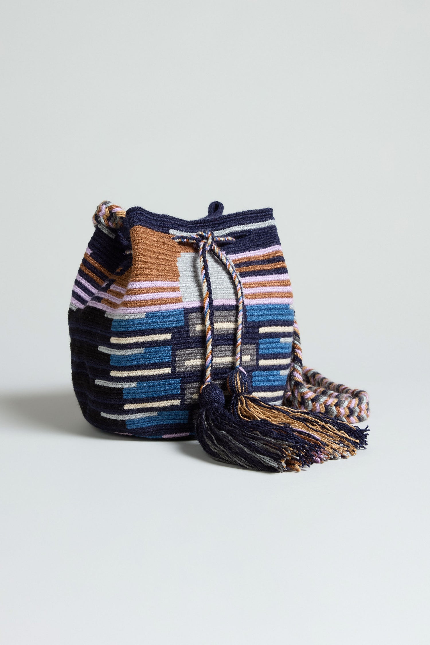 The Handcrafted Crossbody Tassel Bag is a woven bucket-style accessory with a geometric striped pattern in blue, brown, white, and purple, finished with a braided shoulder strap and playful tassel detail.