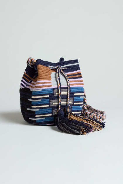 The Handcrafted Crossbody Tassel Bag is a woven bucket-style accessory with a geometric striped pattern in blue, brown, white, and purple, finished with a braided shoulder strap and playful tassel detail.