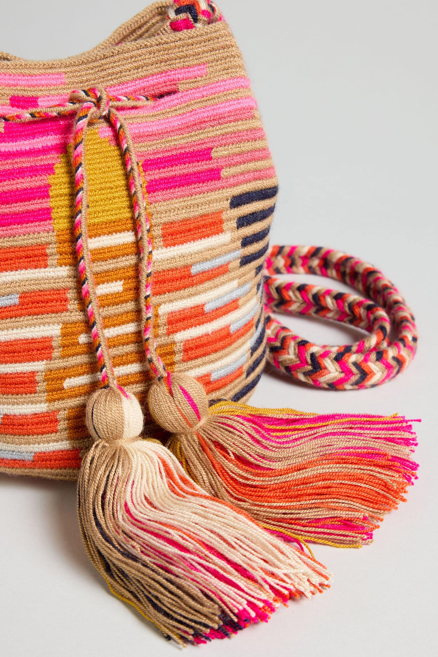 The Handcrafted Crossbody Tassel Bag features tasseled drawstrings, a matching braided strap, and chic boho stripes in pink, orange, beige, and yellow.
