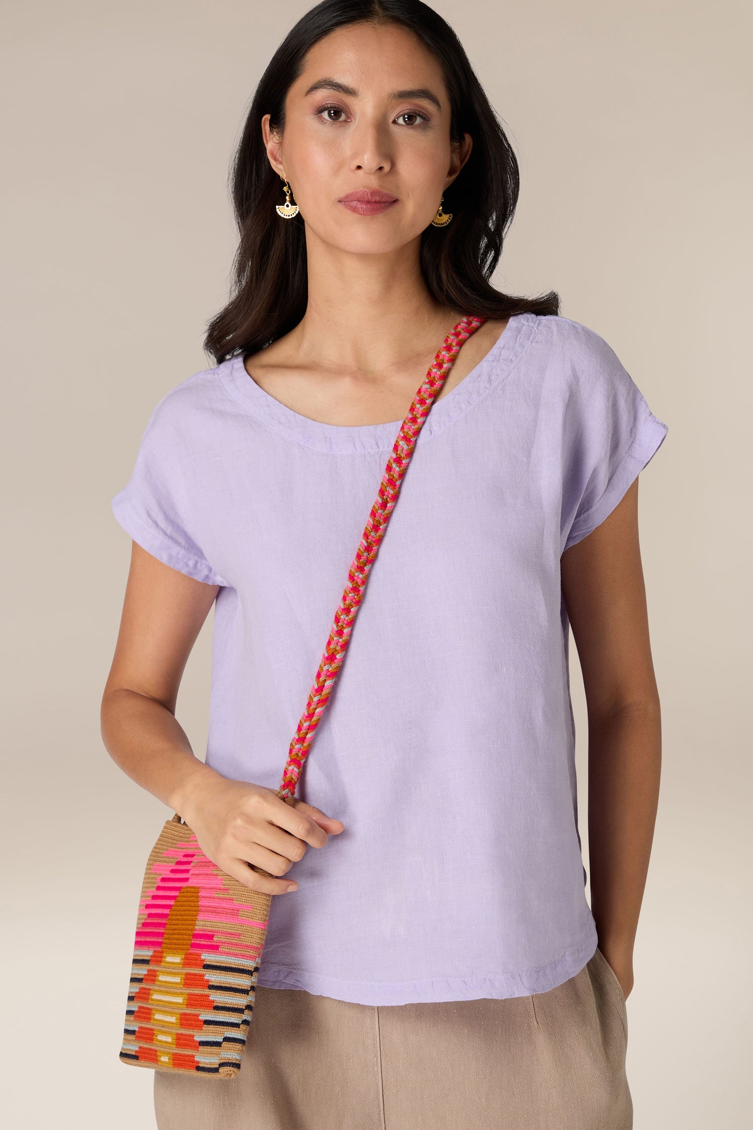 A woman with long dark hair wears a light purple top and beige pants, holding a vibrant Handcrafted Rectangular Crossbody Bag with a red strap over her shoulder. She has gold earrings and a neutral expression.