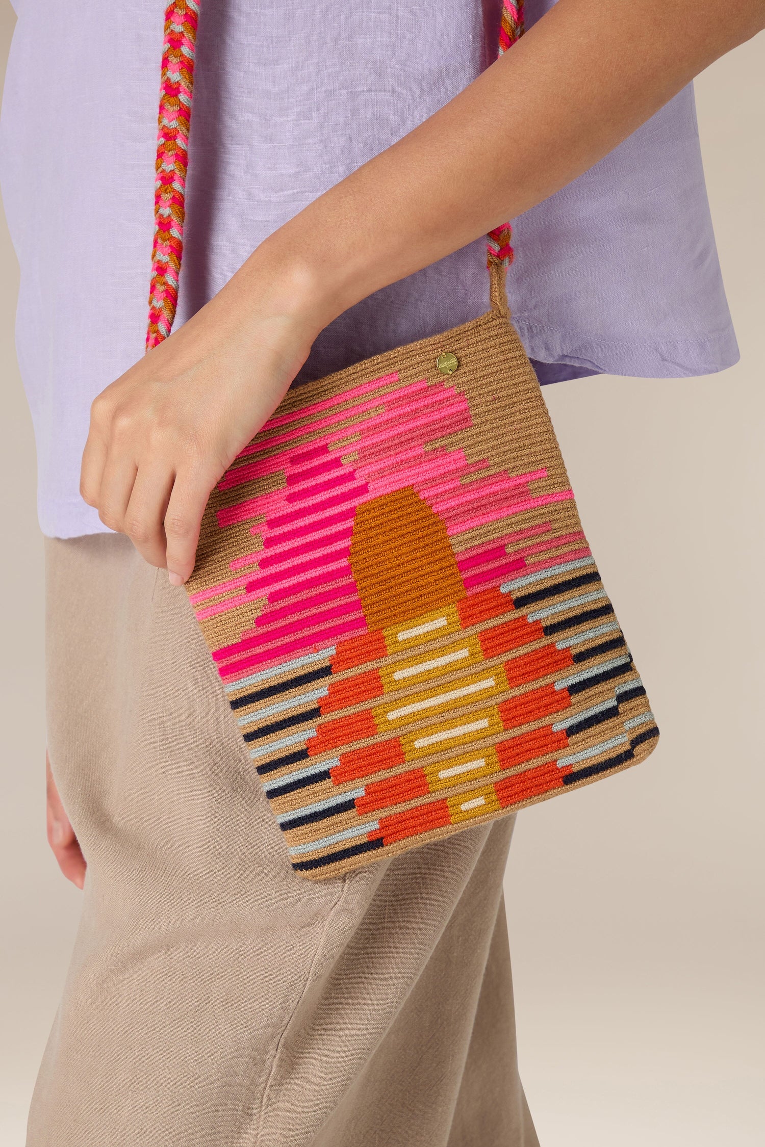 Person holding a small, vibrant Handcrafted Rectangular Crossbody Bag with an abstract pink, orange, and yellow design, wearing a light purple top and beige pants. The traditional techniques used in its creation add to its charm.