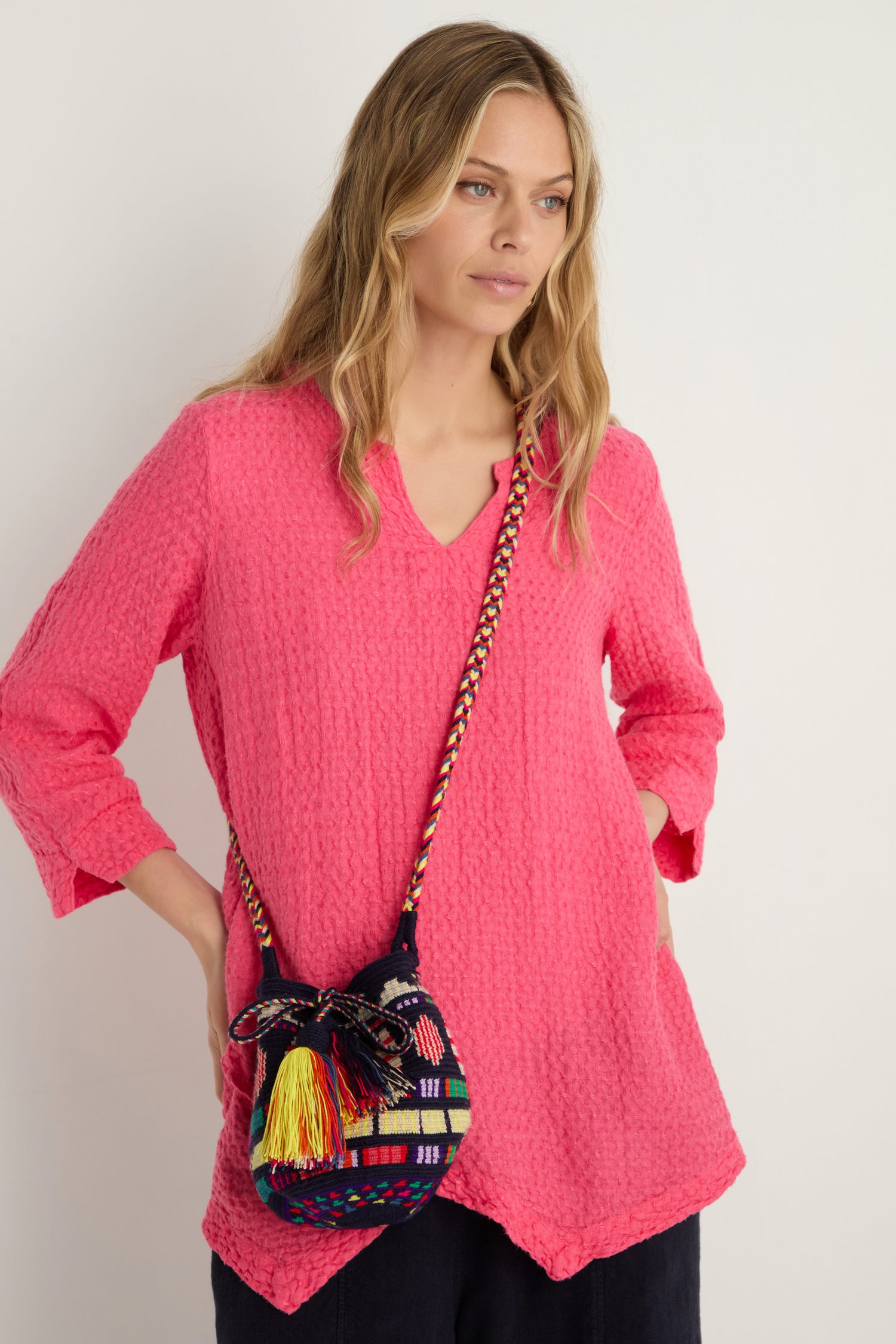 A woman in a textured bright pink tunic holds the Hand Crocheted Wayuu Geometric Crossbody Bag with tassels and a braided strap against a plain white background.