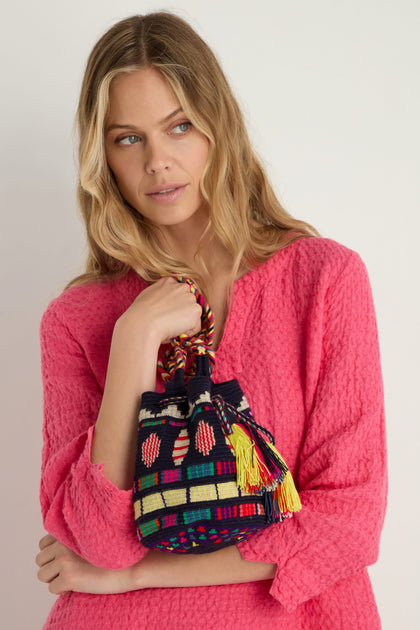A woman in a textured pink top holds the Hand Crocheted Wayuu Geometric Crossbody Bag, featuring colorful patterns, tassels, and a braided handle, against a plain background.
