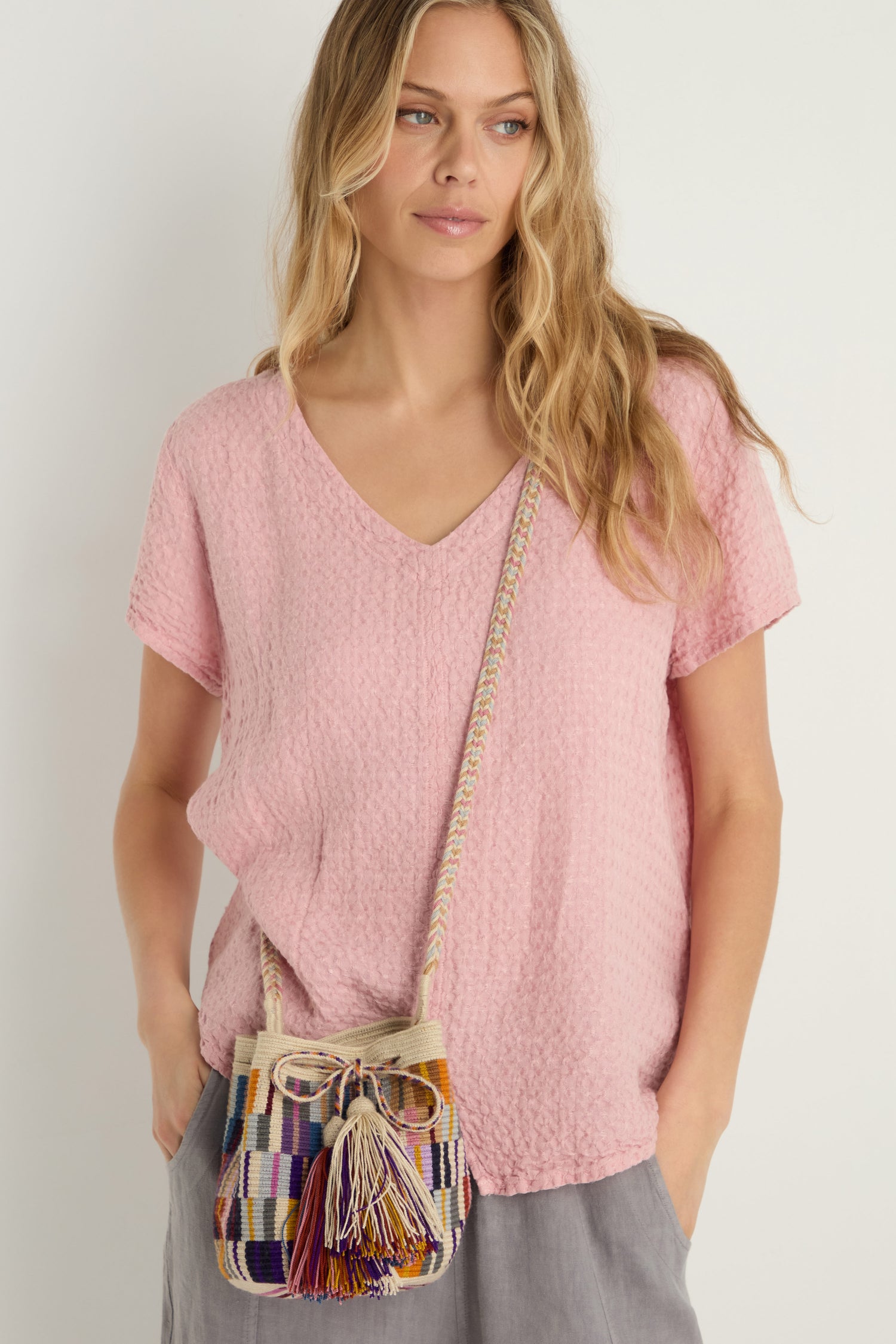 Woman wearing a textured pink short-sleeve blouse and light gray pants, holding a Hand Crocheted Wayuu Crossbody Bag with multicoloured stripes and colorful tassels.