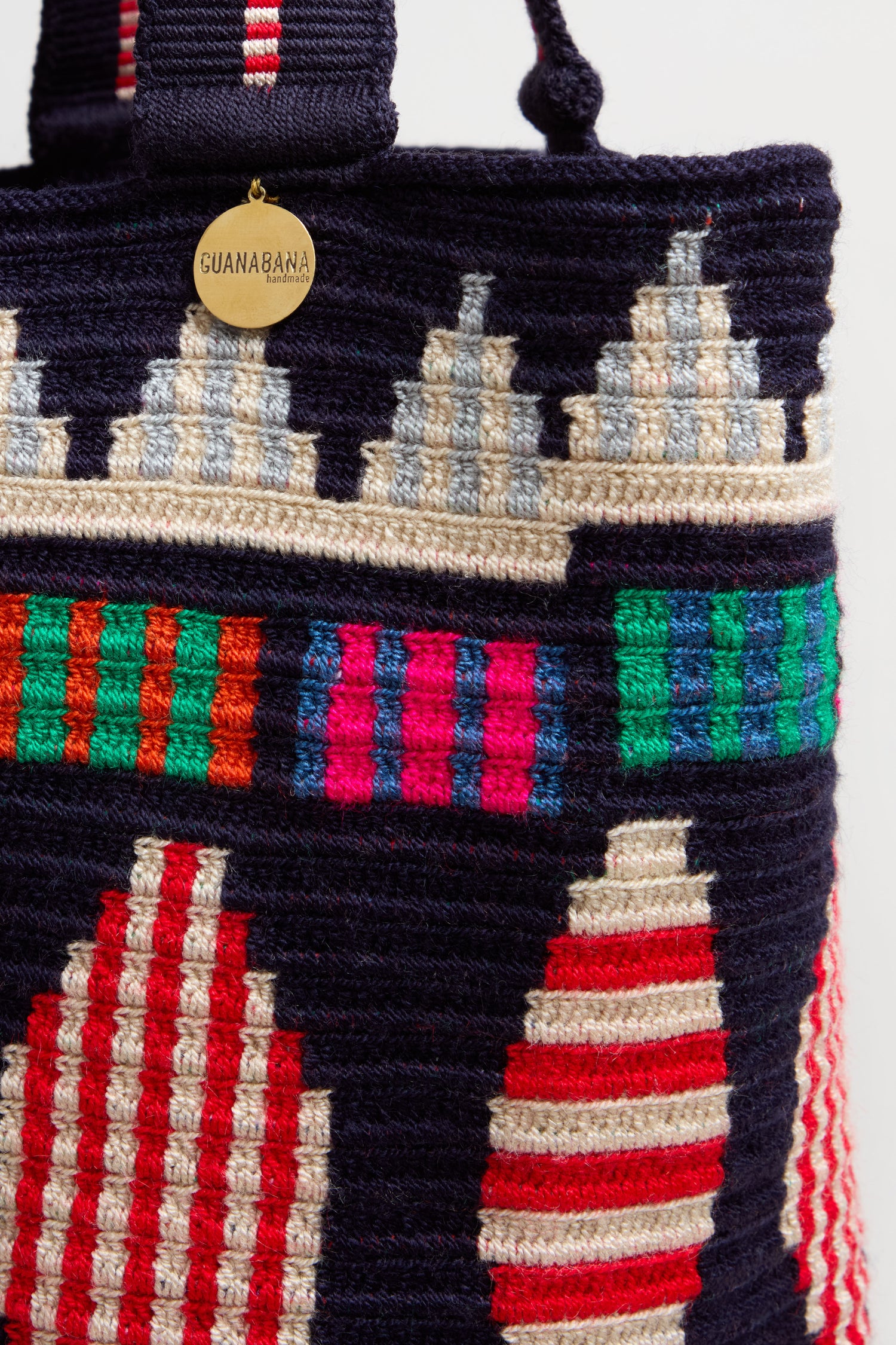 Close-up of the Hand Crocheted Cala Geometric Tote Bag from Colombia, displaying red, green, blue, and cream geometric patterns and a round gold tag labeled "Guanabana.