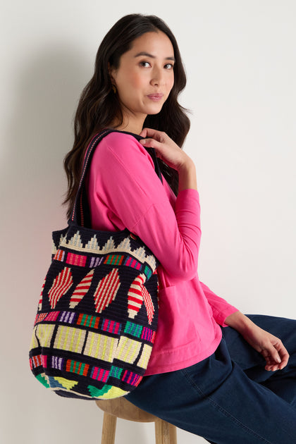 A woman sits on a stool in a bright pink top and blue pants, carrying the Hand Crocheted Cala Geometric Tote Bag, featuring bold Colombian-inspired patterns, over her shoulder.