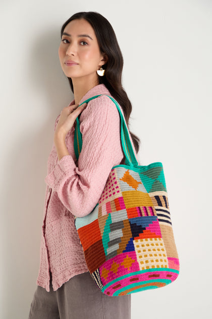 A woman stands against a plain light background, wearing a textured pink blouse and gray pants, carrying the Hand Crocheted Cala Tote Bag with a colorful geometric design over her shoulder.