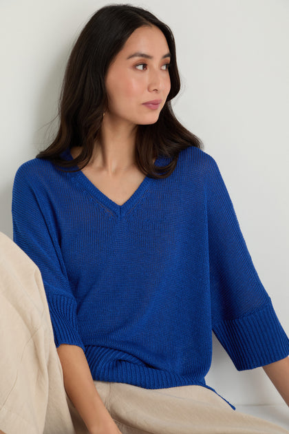 Woman with long dark hair wears a blue V Neck Relaxed Knit Top and light beige pants, sitting and looking to the side against a plain background.