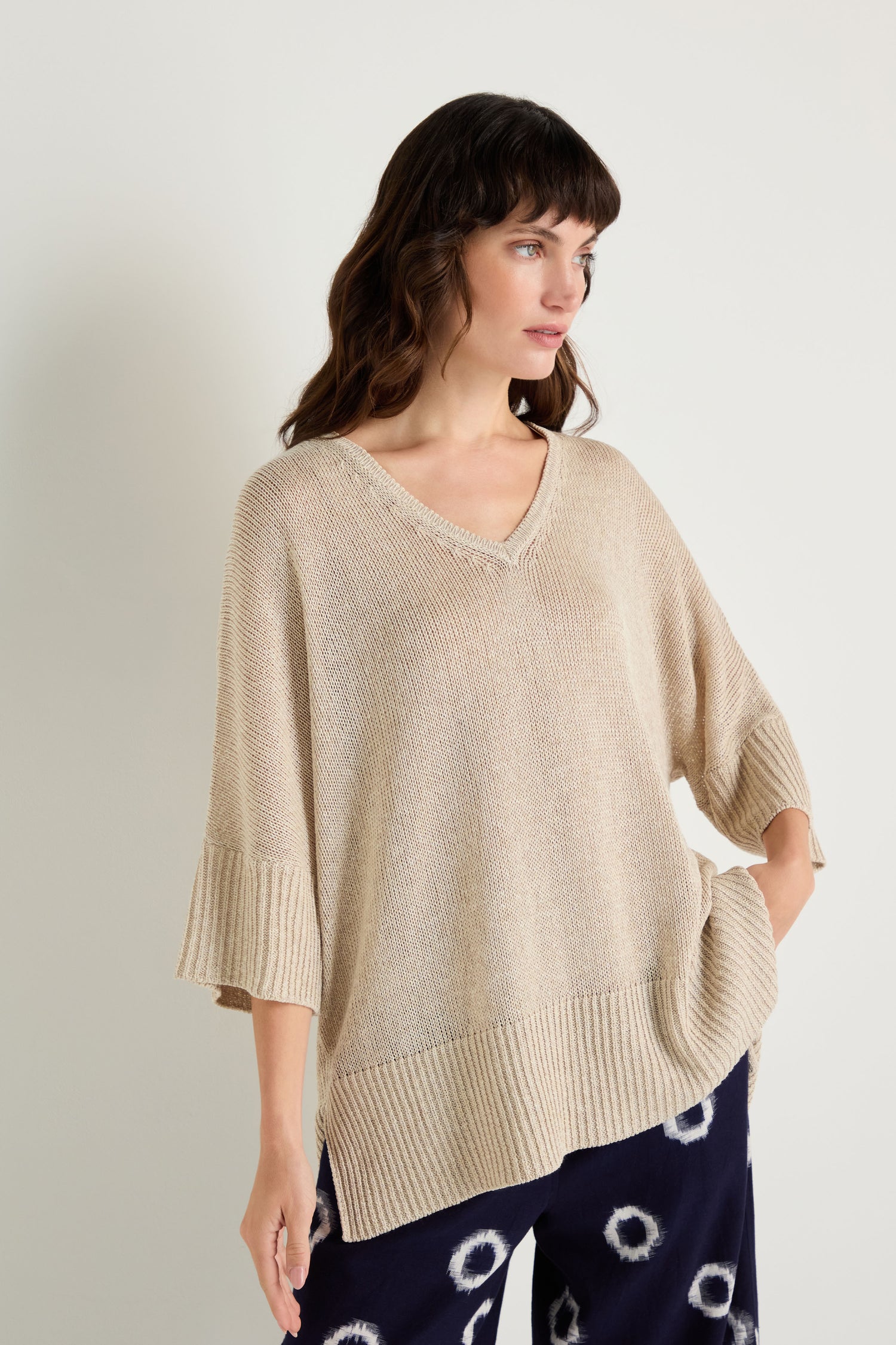 A woman wears the V Neck Relaxed Knit Putty Top with wide sleeves and dark pants featuring white circular patterns, standing against a plain light background.