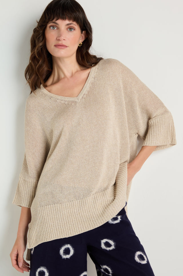 A woman wearing a V Neck Relaxed Knit Putty Top with elbow-length sleeves and navy pants featuring a white circular pattern stands against a plain background.