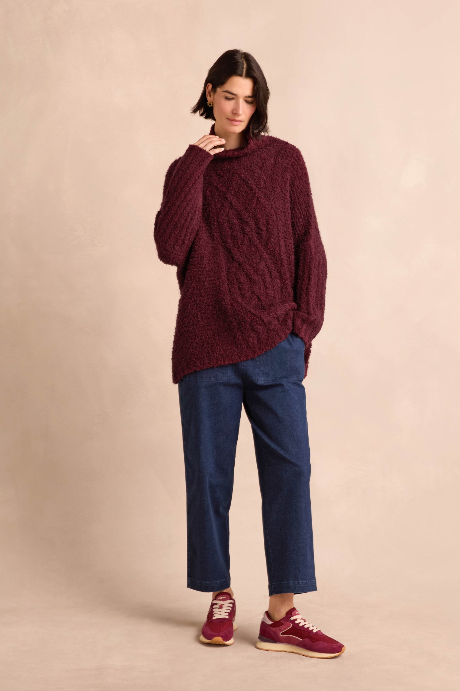 A woman stands against a beige background wearing the Bordeaux Boucle Cable Knit Jumper, paired with blue jeans and matching burgundy sneakers, with one hand touching her chin and the other tucked into her pocket.